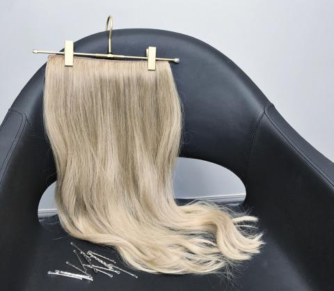 Real Hair Clip In Extensions |A blonde clip on hair extension hanging from a golden hanger. They've both been placed on top of a black leather salon chair. 